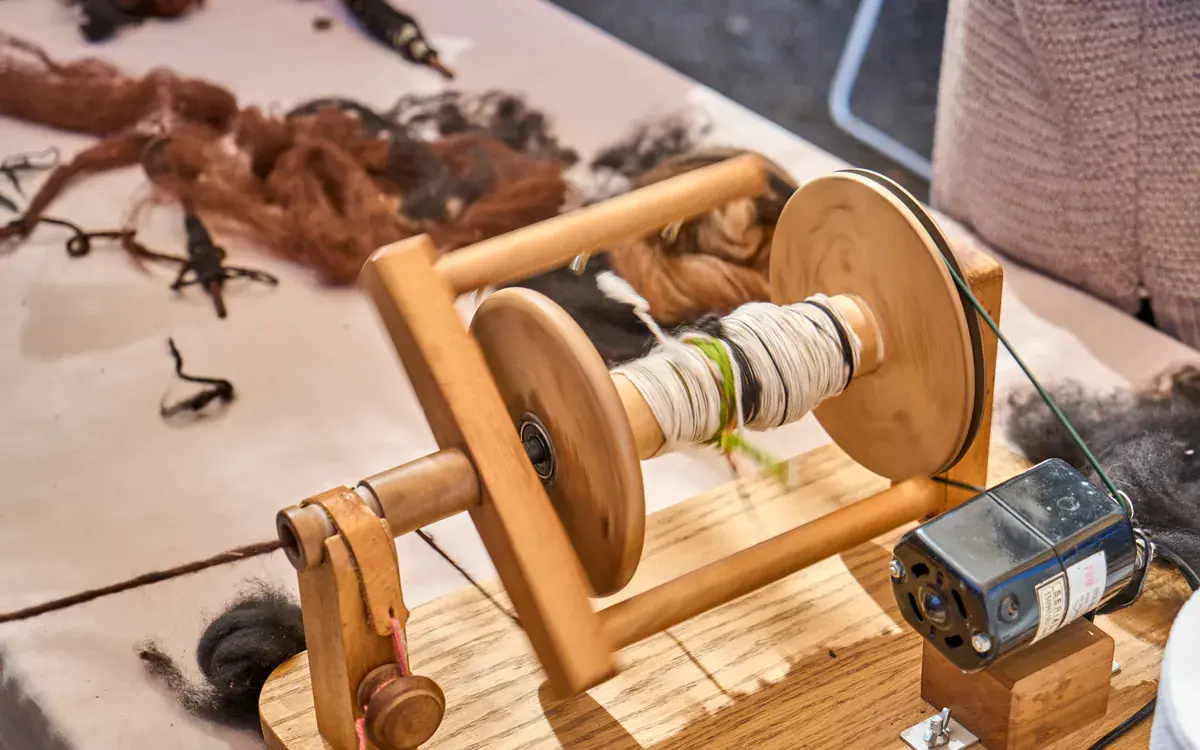 A wooden spinning wheel turns, winding white and black yarn, with brown fibre on the table