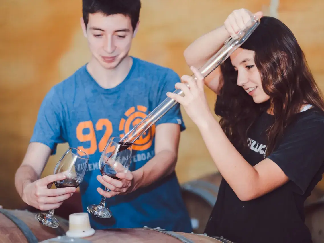 Two young visitors draw wine from the barrel with a glass pipette and pour it into a glass