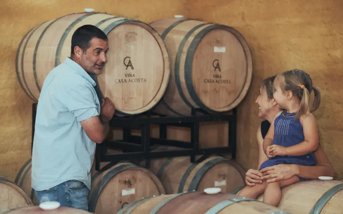 A visitor stands by barrels bearing the Viña Casa Acosta logo while a woman and a little girl sit on another barrel