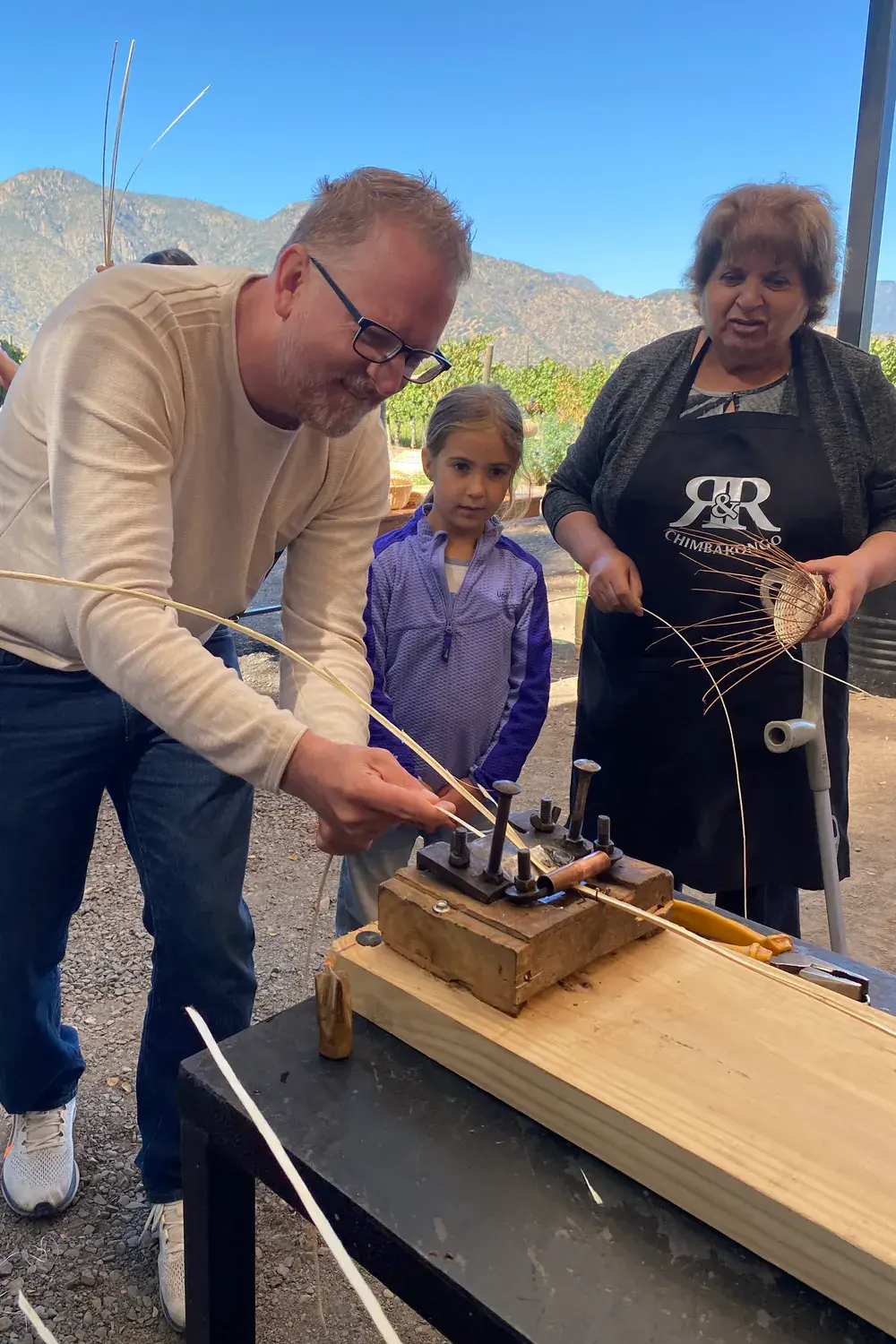 A participant prepares a strip of wicker in the cutting tool, guided by an artisan, with the vineyard and the mountains behind