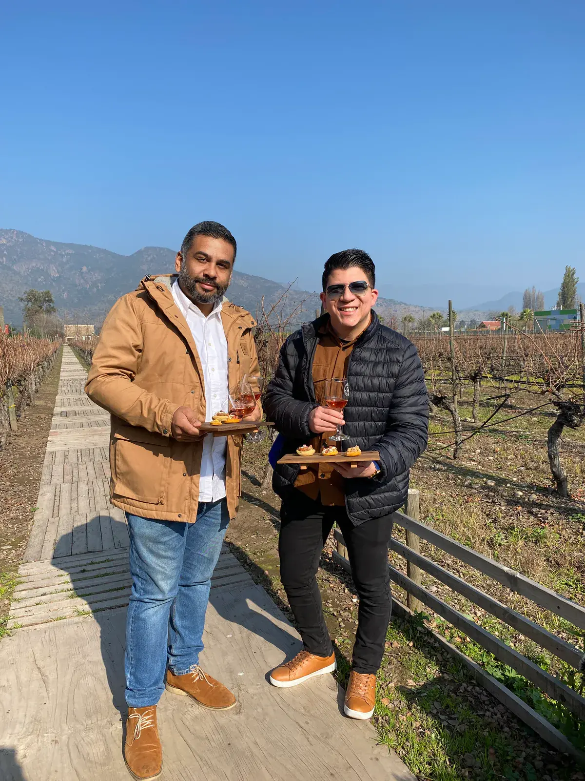 Two visitors smile with glasses of rosé and boards of bites on a wooden walkway among the vines
