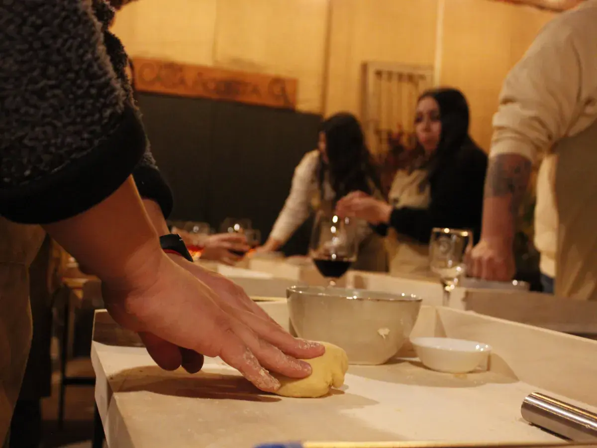 A hand kneads a small ball of dough on the board, with wine glasses and participants behind