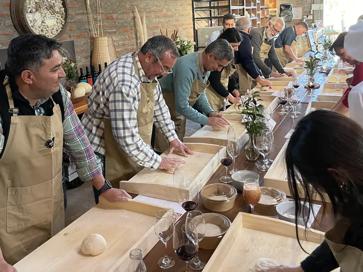 Participants in aprons knead by hand on wooden trays, with wine glasses on the table