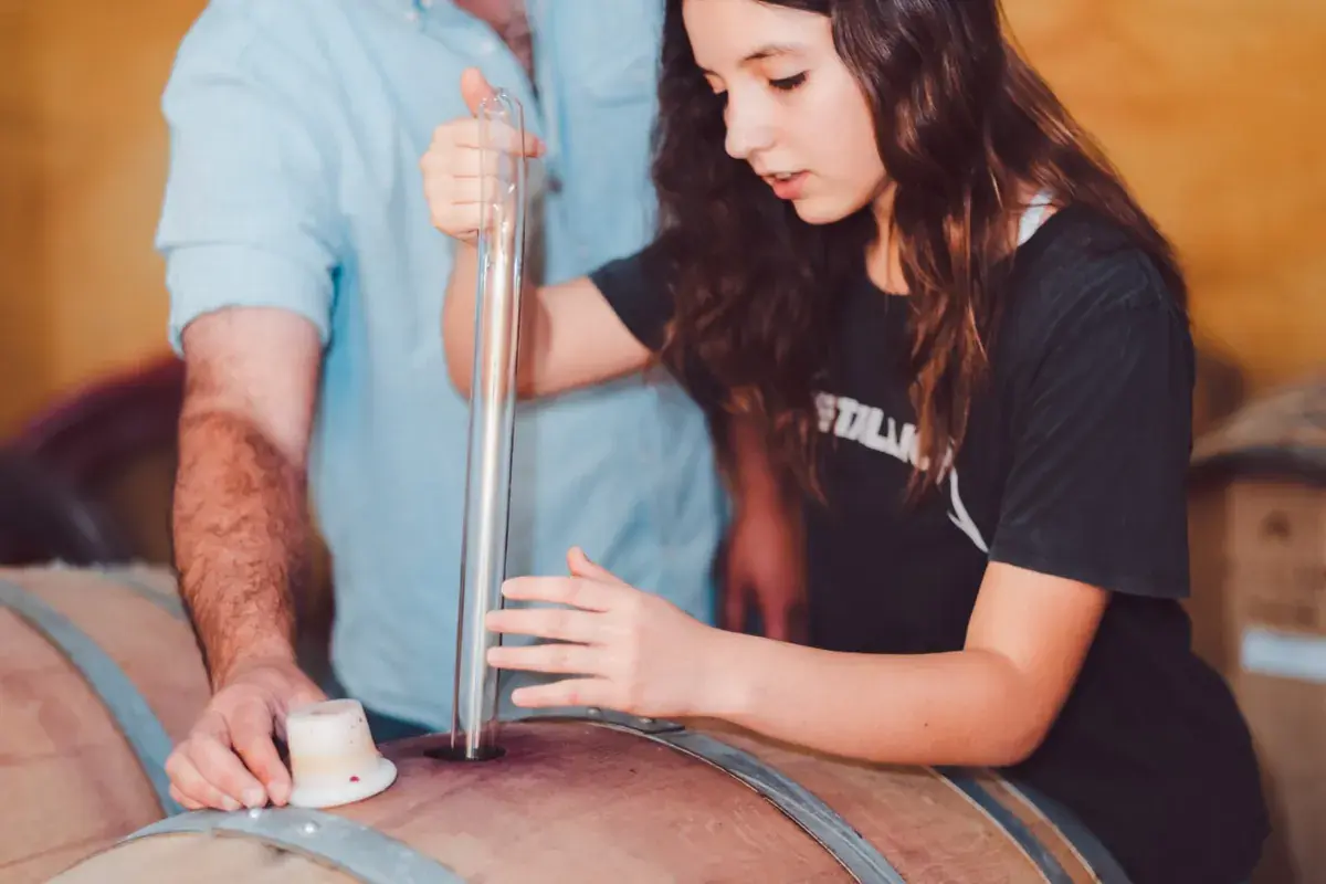 A young visitor draws wine from the barrel with a glass pipette, next to another guest