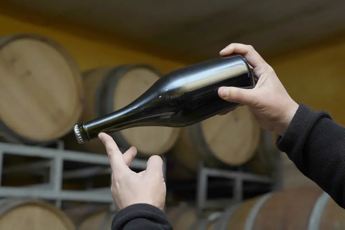 Hands hold a bottle of wine in front of the cellar's barrels