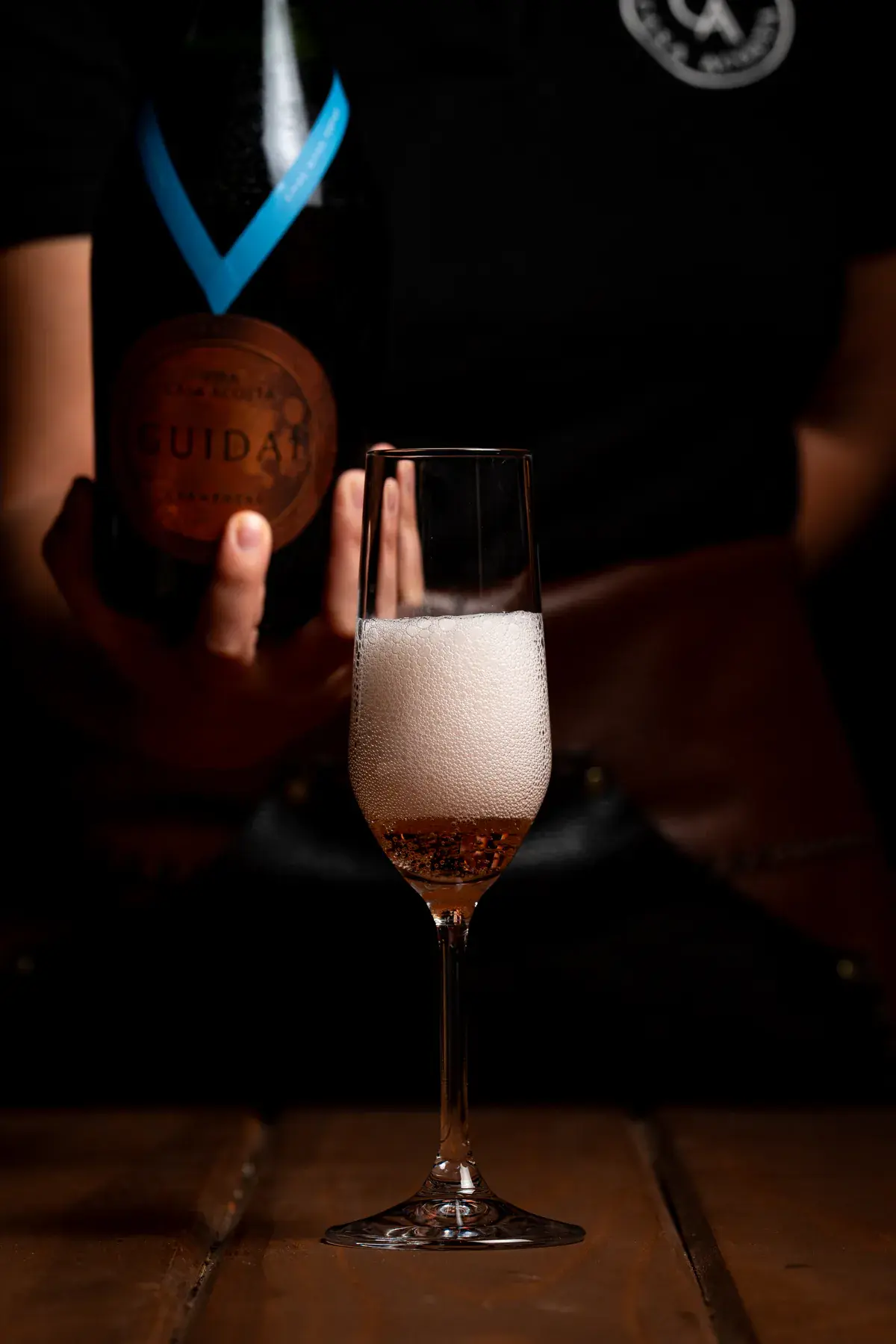 A bottle of Guidaí sparkling wine and a flute with pink foam, against a dark background