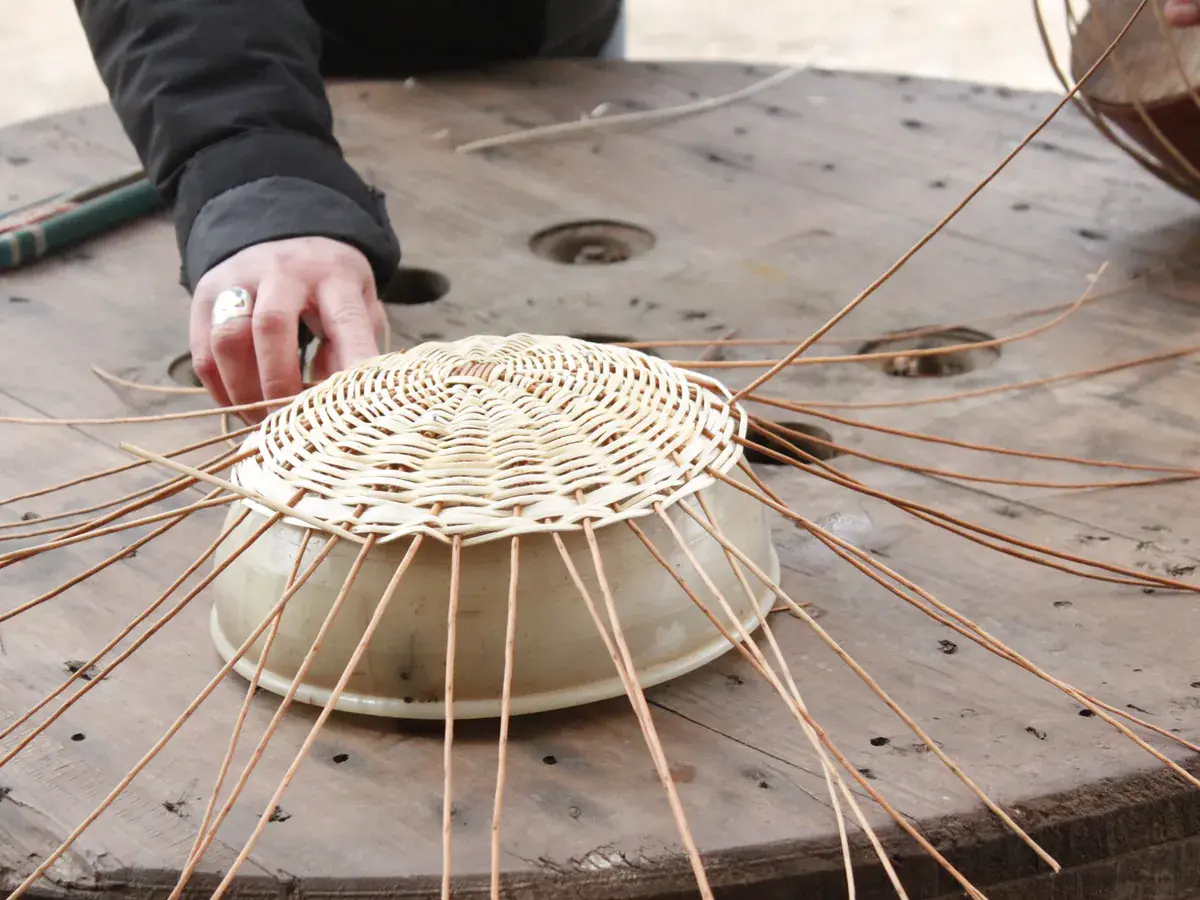 Manos tejiendo la base de una pieza de mimbre sobre un molde, en la mesa del taller