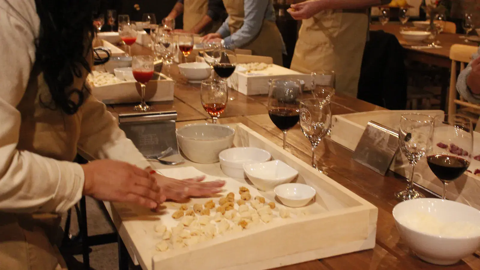 Hands shape ridged gnocchi on a wooden tray beside glasses of red wine