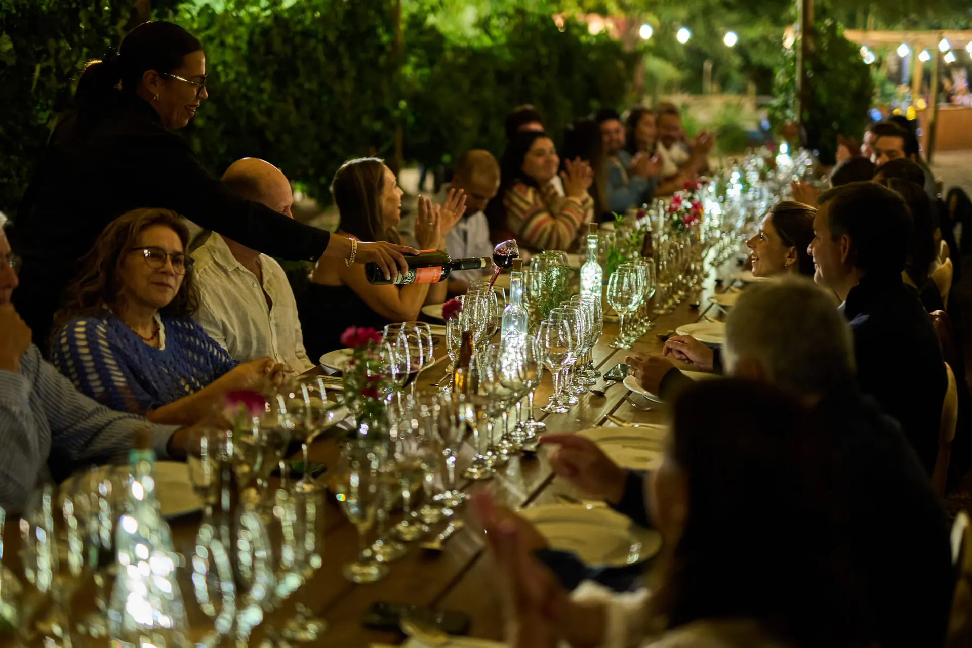 Night-time dinner at a long table among the vines: someone pours red wine while the guests talk